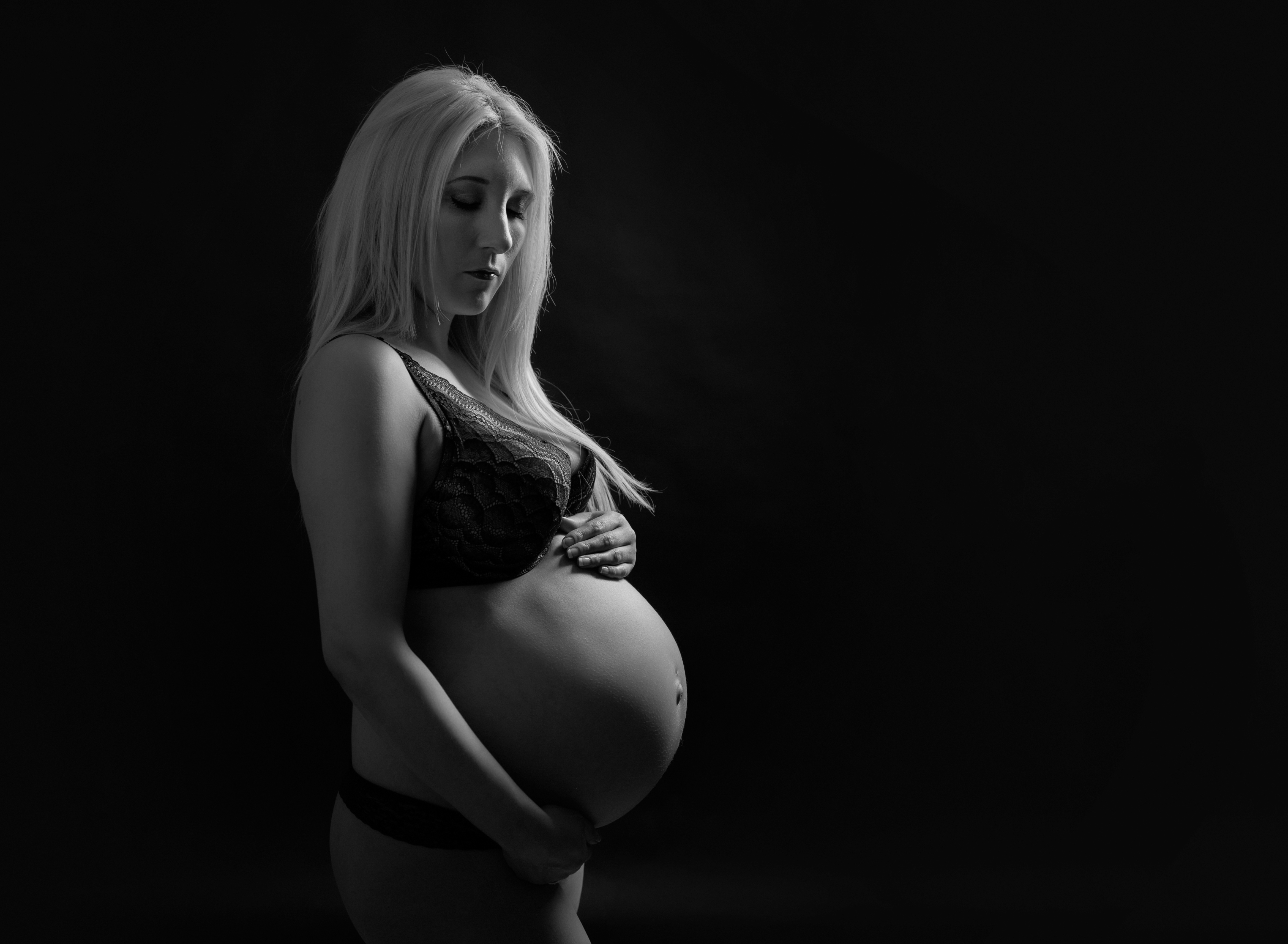 Studio maternity portrait on a dark background, with the pregnant mother standing in a black dress, her expectant belly featured, while she looks at the camera. Image by Helga Himer Photography, Sudbury, Ontario.