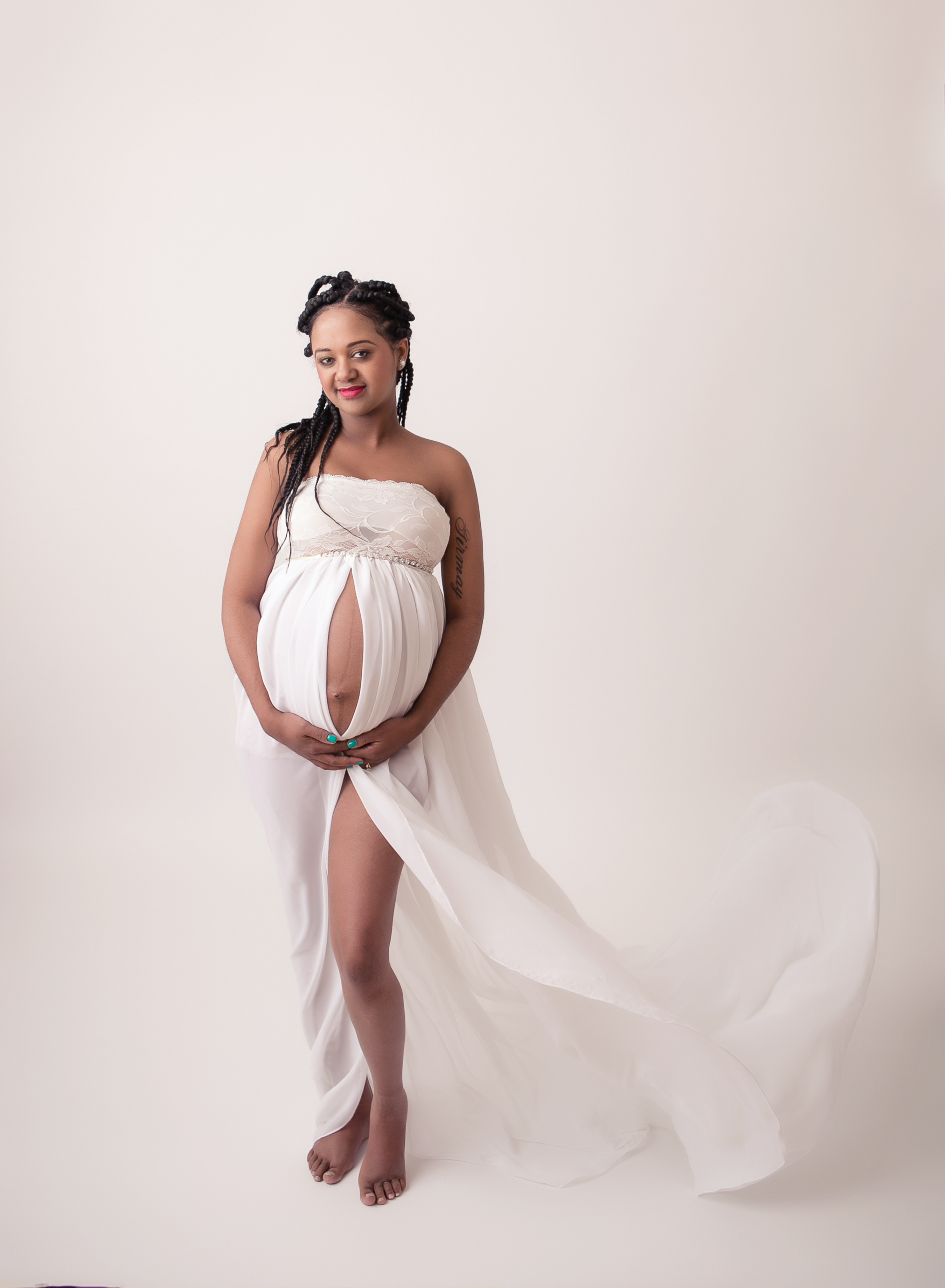 Studio maternity portrait on a light background with the pregnant black mother in a white dress holding around her expectant belly and smiling at the camera. Image by Helga Himer Photography, Sudbury, Ontario.