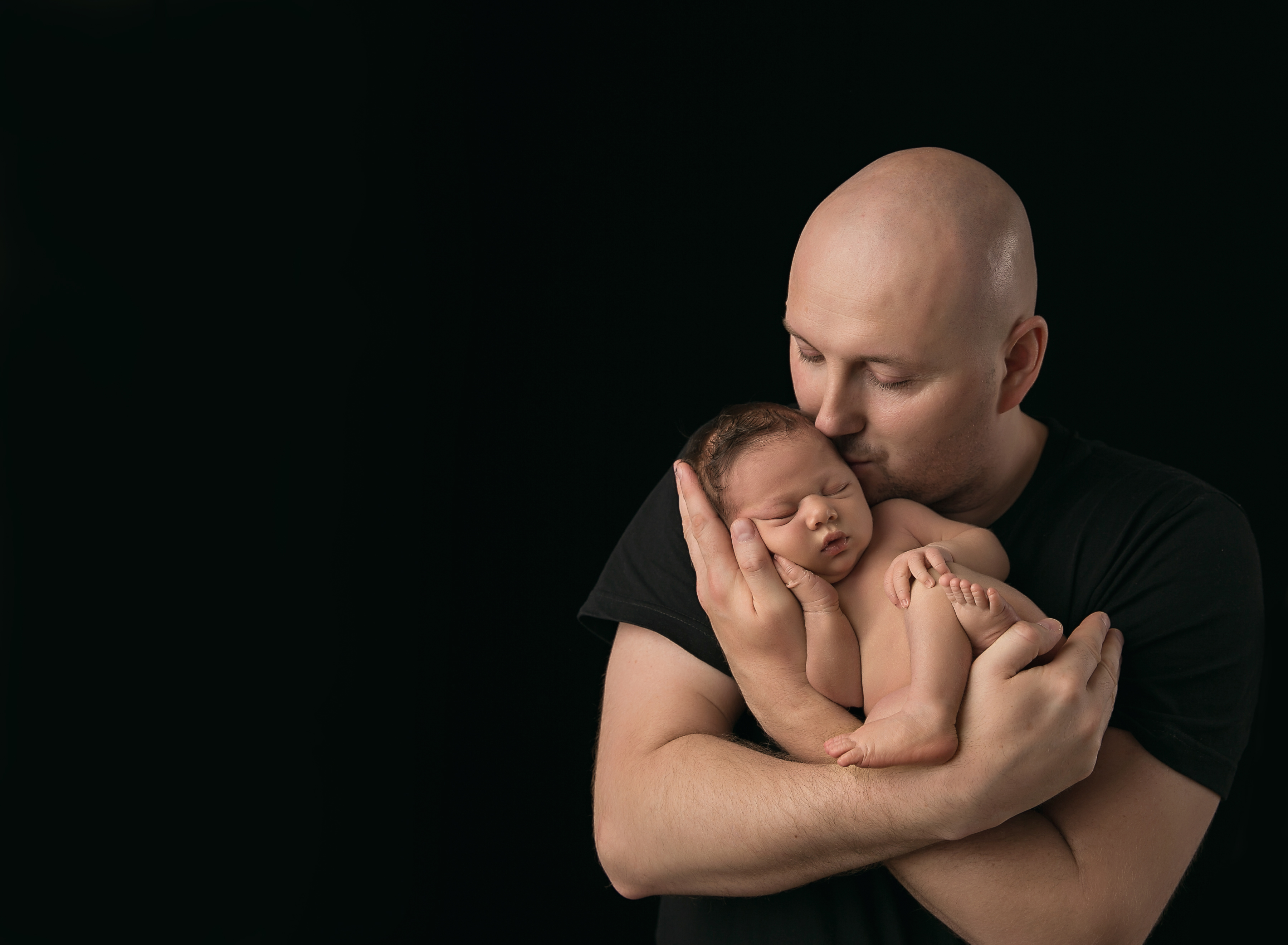 Studio portrait with dark background of an affectionate father holding his newborn sonin his arms. Image by Helga Himer Photography, Sudbury, Ontario.