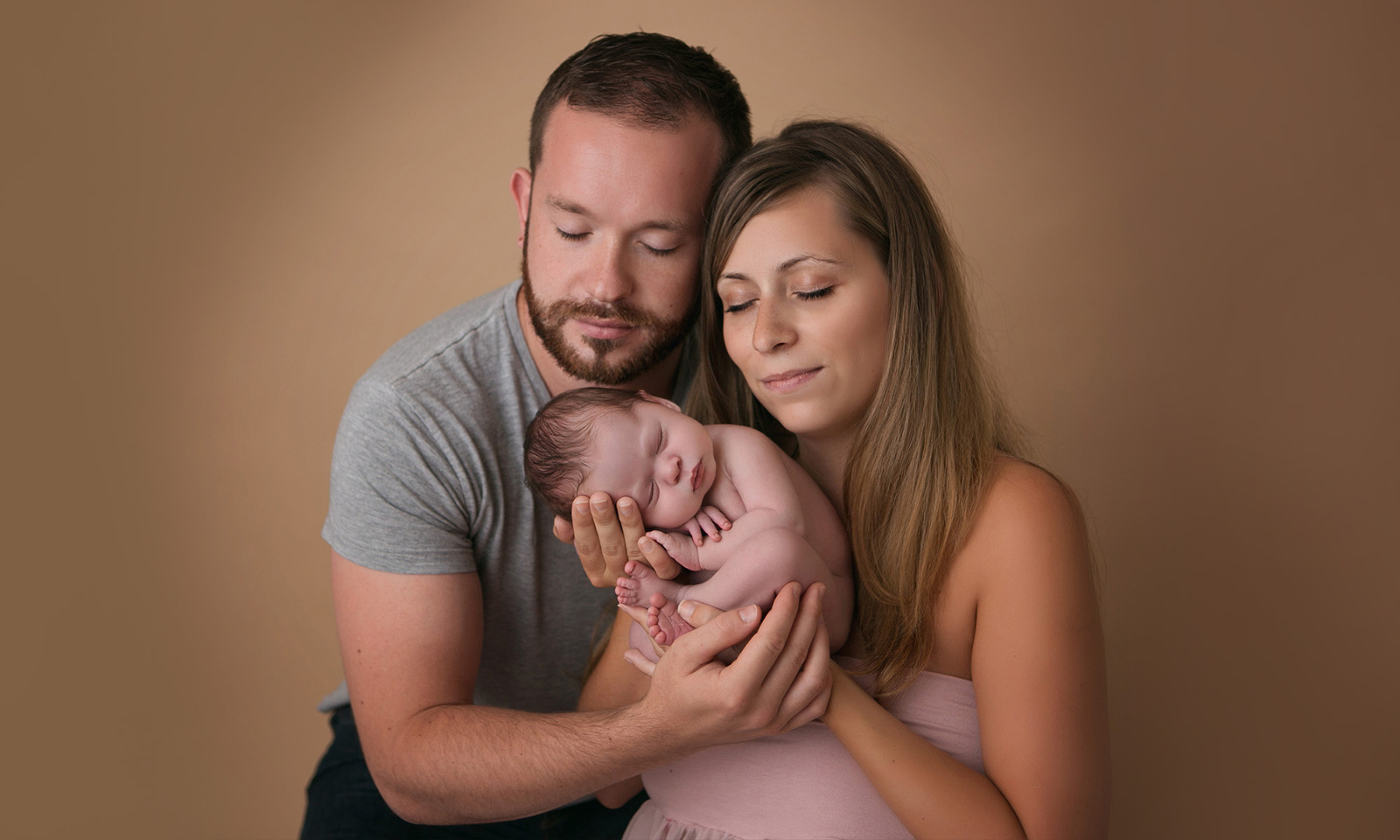 Studio family newborn portrait session with beige background of a mother and father with eyes closed, holding their sleeping newborn son between them. Image by Helga Himer Photography, Sudbury, Ontario.