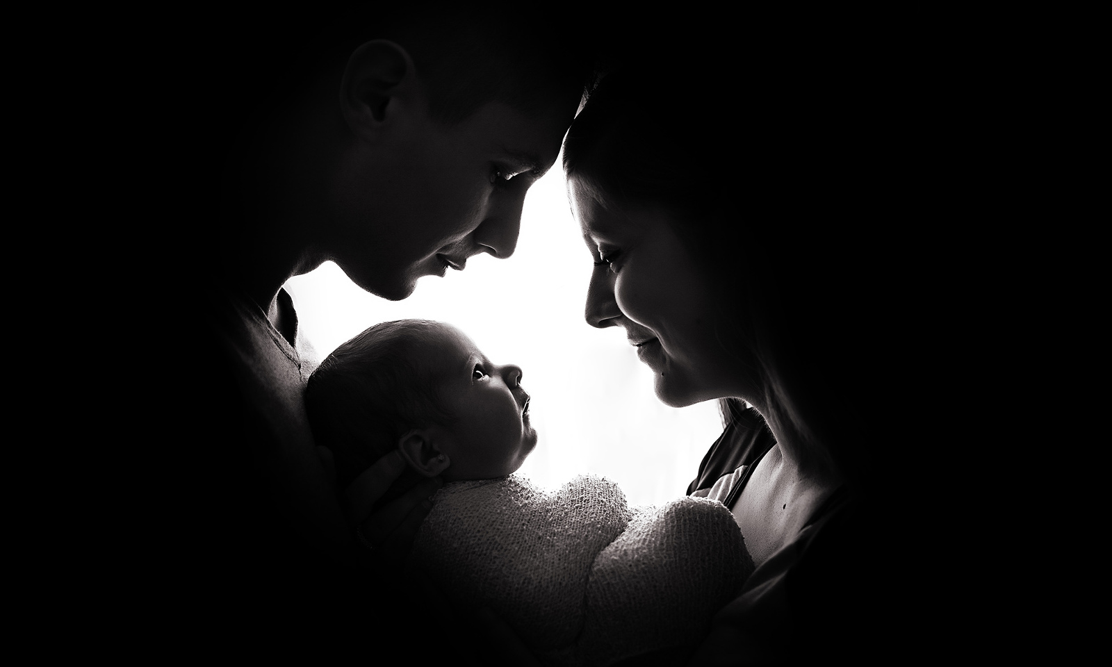 Award-winning black-and-white studio newborn family portrait, from the side, with a mother and father smiling down from above upon their alert, wrapped newborn baby, who is smiling back at them. Image by Helga Himer Photography.