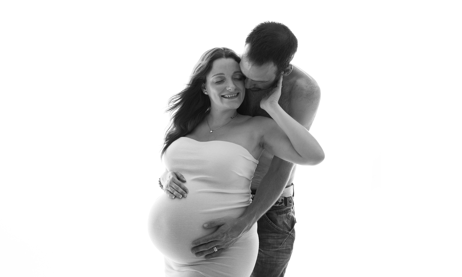 Black and white studio couples maternity portrait featuring a white background and taken from the front, with the pregnant mother in a white dress, and the shirtless father's are wrapped around her expectant belly from the back. Image by Helga Himer Photography, Sudbury, Ontario.