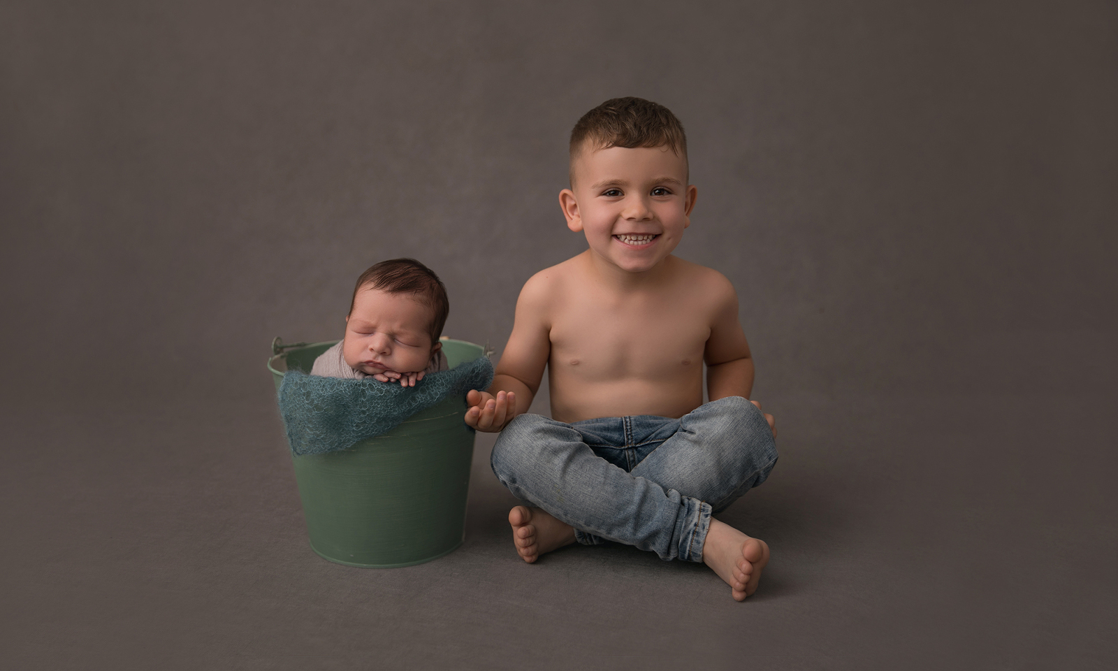 Studio newborn portrait session from the front with grey background of a proud older toddler brother sitting next to his newborn sibling is is sleeping in a green pail. Image by Helga Himer Photography, Sudbury, Ontario.