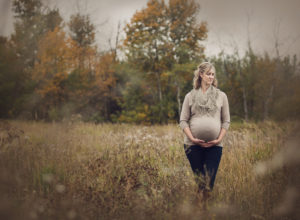 Outdoor fall maternity portrait in a grassy field, the pregnant mother standing in grey shirt and dark pants, looking in the distance, holding her expectant belly. Image by Helga Himer Photography, Sudbury, Ontario.