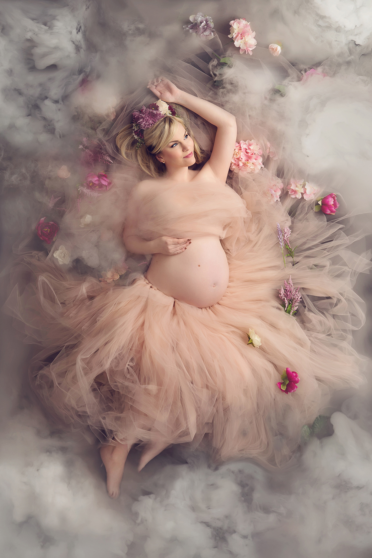 Studio maternity dry ice portrait from above, with the pregnant mother lying down, her light pink dress spread around her, looking off to the distance, surrounded by fog and mist, her expectant belly exposed. Image by Helga Himer Photography, Sudbury, Ontario.