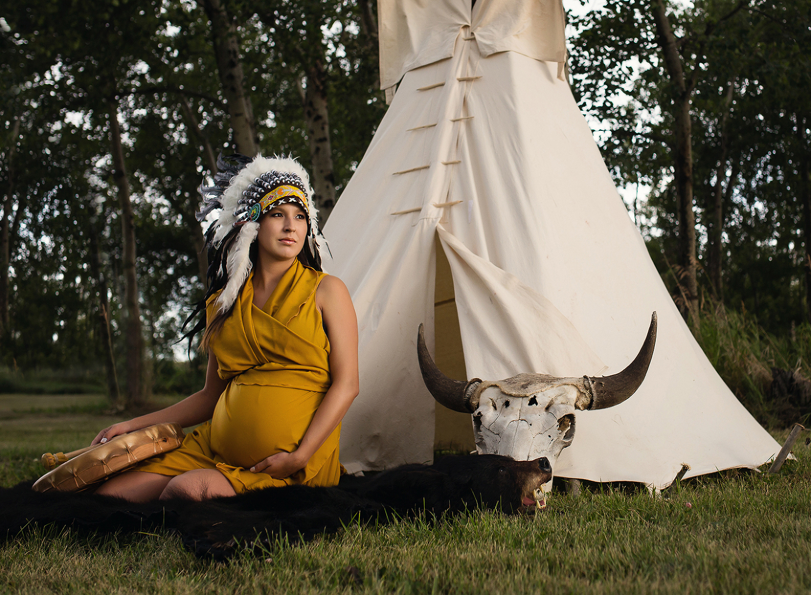 Outdoor maternity portrait in a forest, the pregnant Indigenous mother is wearing a native Indian headdress and is sitting in front of a beige tipi in an elegant yellow dress at sunset, looking off into the distance, an arm around her expectant belly, by the entrance to the wigwam. Image by Helga Himer Photography, Sudbury, Ontario.