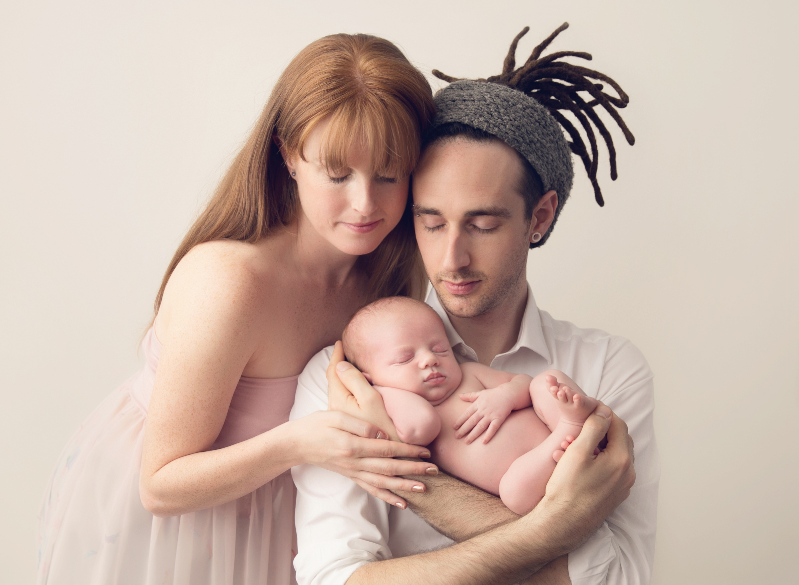 Studio portrait with light background of a happy mother and father, smiling together with their sleeping newborn baby in the mother's arms. Image by Helga Himer Photography, Sudbury, Ontario.