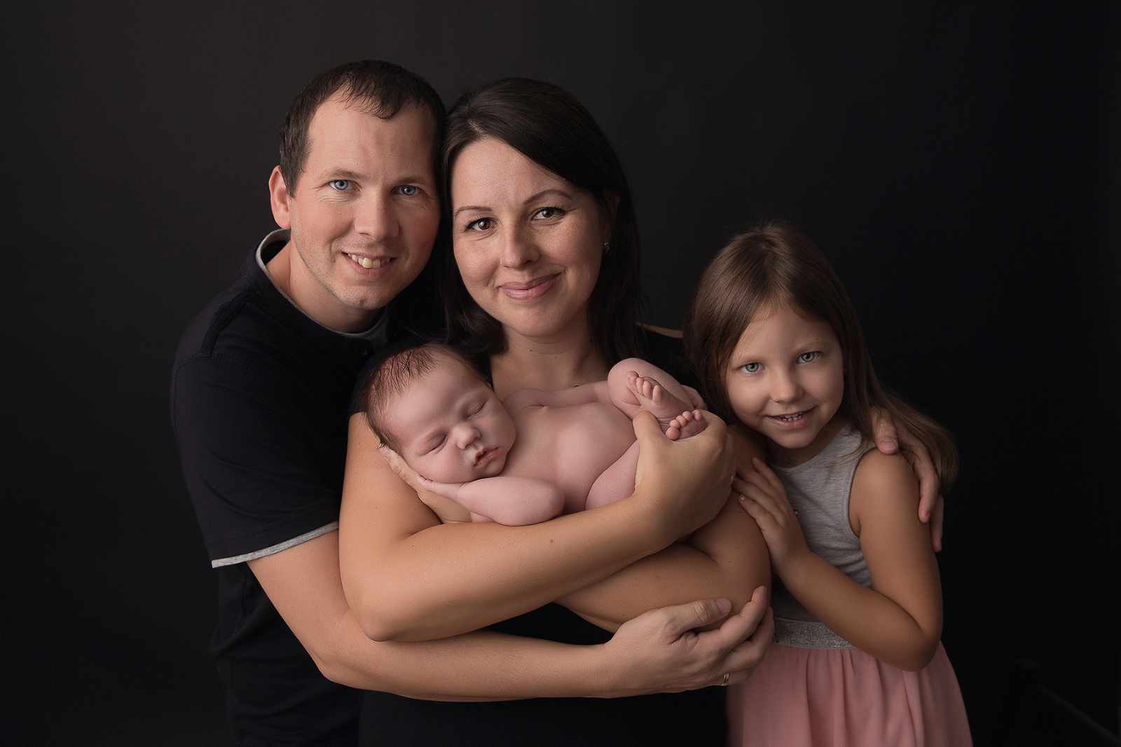 Studio family portrait with dark background of a happy family, mother and father and young daughter, smiling together with their sleeping six-day-old newborn baby in the mother's arms. Image by Helga Himer Photography, Sudbury, Ontario.