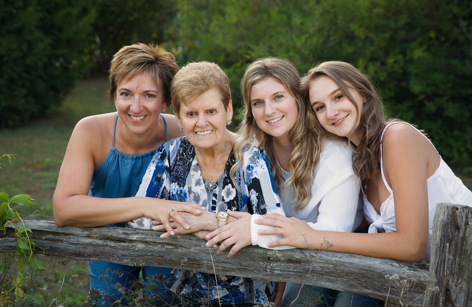 Outdoor portrait session featuring a generational photo with grandmother, mother, and two daughters leaning against a rustic fence in Mallard's Landing park in Sudbury, Ontario. Image by Helga Himer Photography.