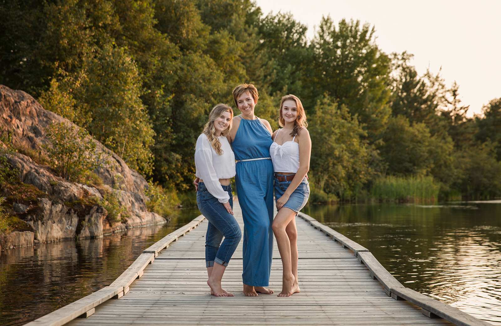 Outdoor portrait session featuring mother and two daughters standing on the dock path in Mallard's Landing park in Sudbury, Ontario. Image by Helga Himer Photography.