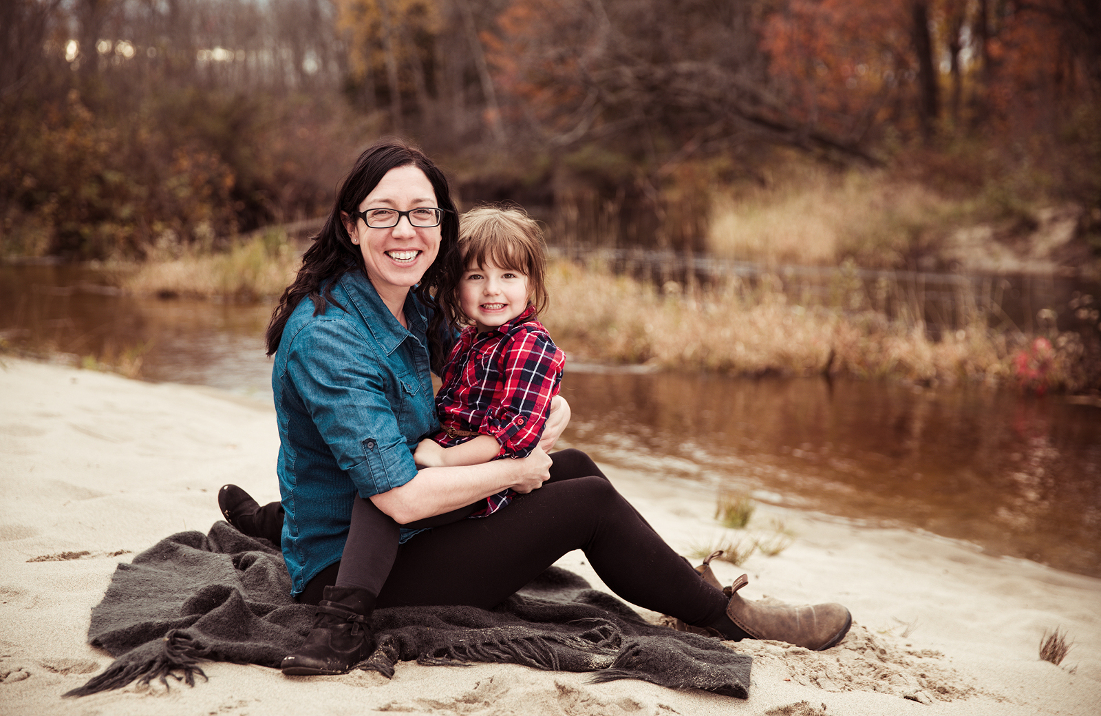 Mother and daughter outdoor portrait session, featuring them sitting together on a sandy beach area of the Whitson River in Val Caron near Sudbury, Ontario on a fall day. Image by Helga Himer Photography.