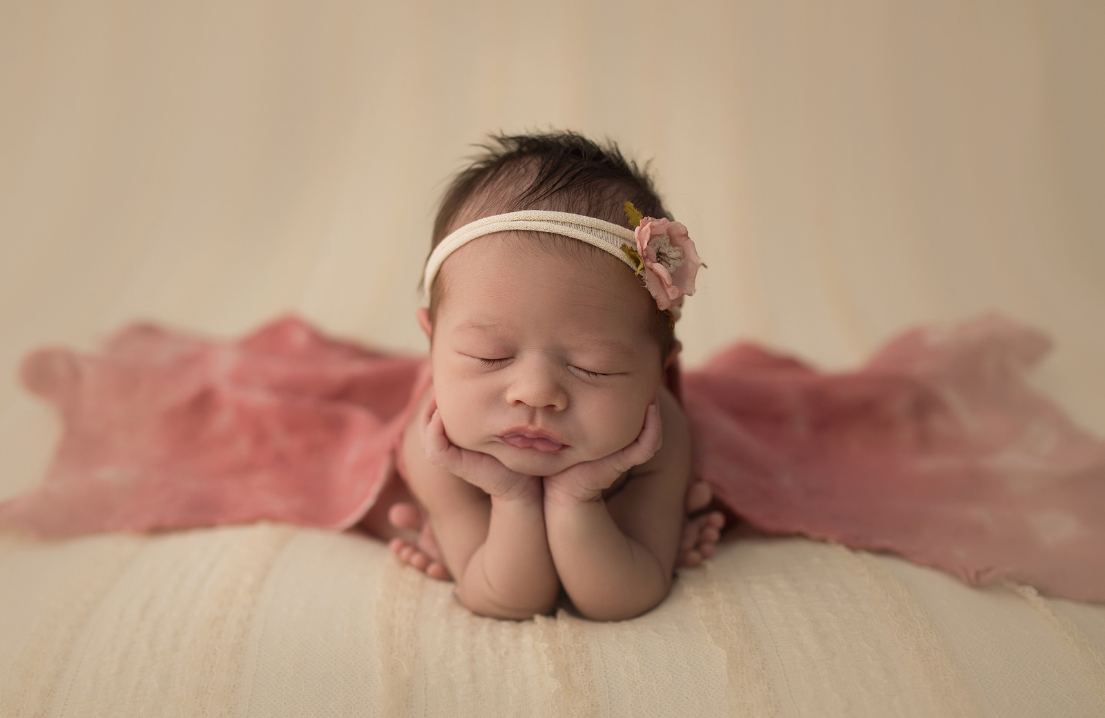 Studio newborn portrait session with cream coloured background from the front, the baby sleeping on a cream-coloured blanket in a froggy pose, along with a flower headband and a light pink blanket behind. Image by Helga Himer Photography, Sudbury, Ontario.