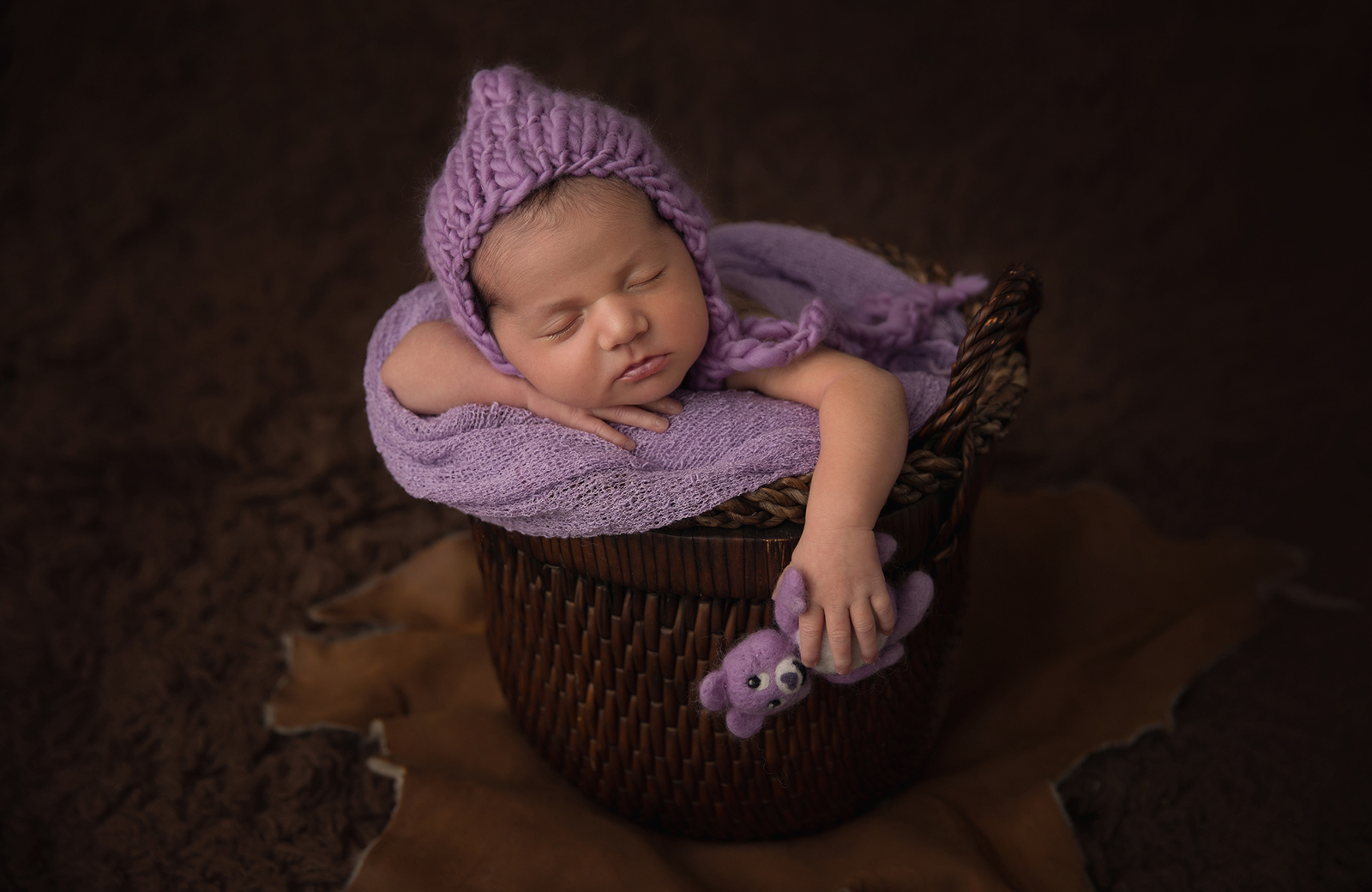 Studio newborn portrait session with dark background, featuring a shot from the side of a sleeping baby in a basket with purple blanket wearing a purple bonnet, and holding onto a small felted purple teddy bear. Image by Helga Himer Photography, Sudbury, Ontario.