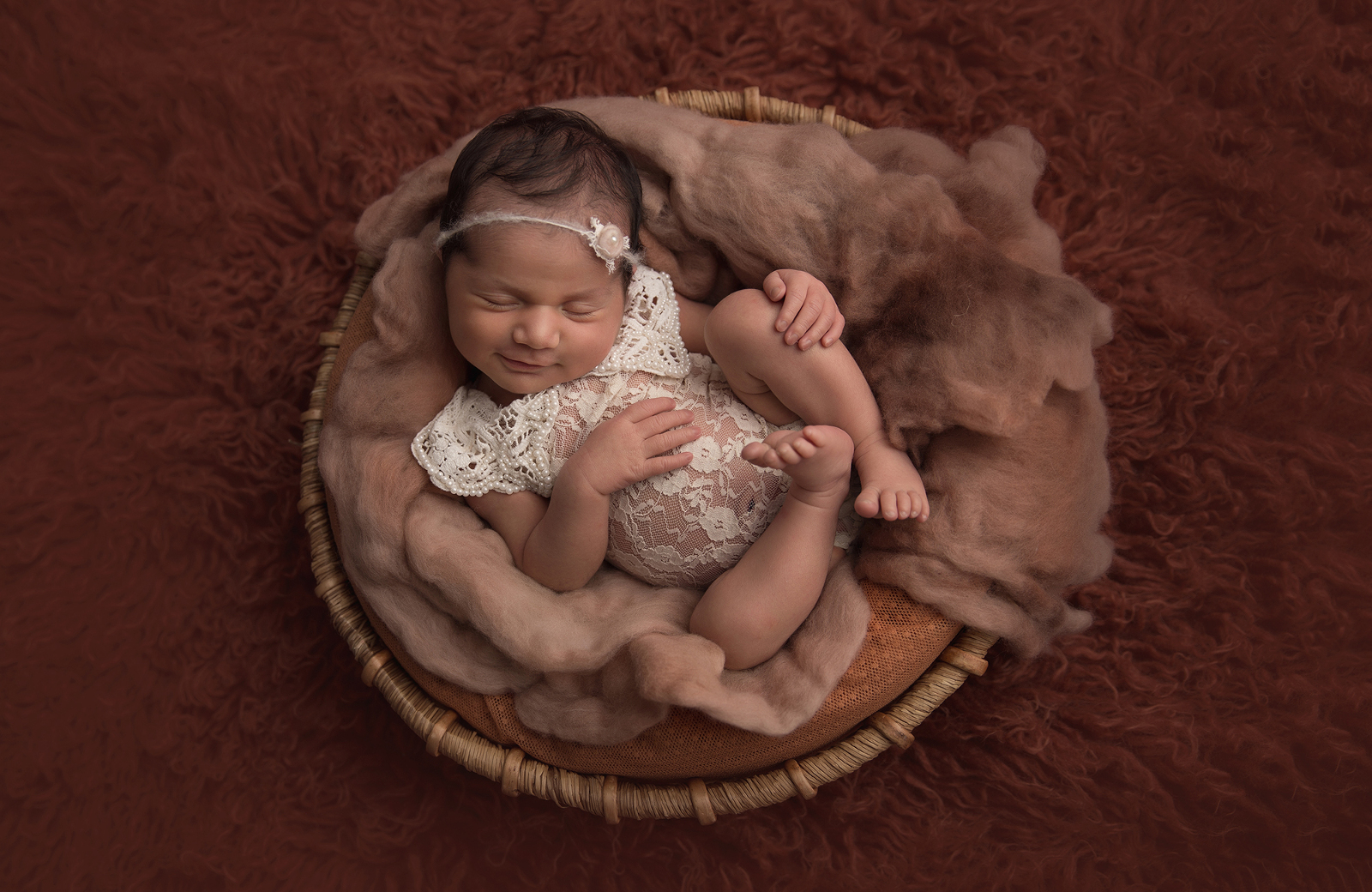 Studio newborn portrait session with brown background, featuring a shot from above of a smiling baby sleeping in a round basket wearing a lace onesie and flower headband. Image by Helga Himer Photography, Sudbury, Ontario.