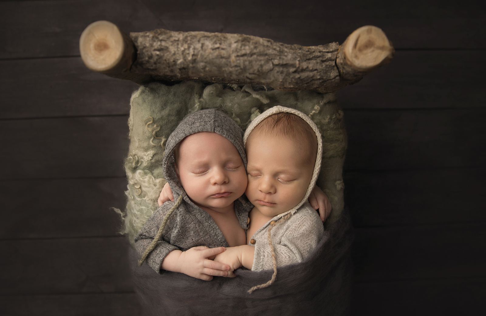 Studio newborn twins portrait session with dark background, the twins sleeping together and holding hands on a custom-made wooden log bed. Image by Helga Himer Photography, Sudbury, Ontario.
