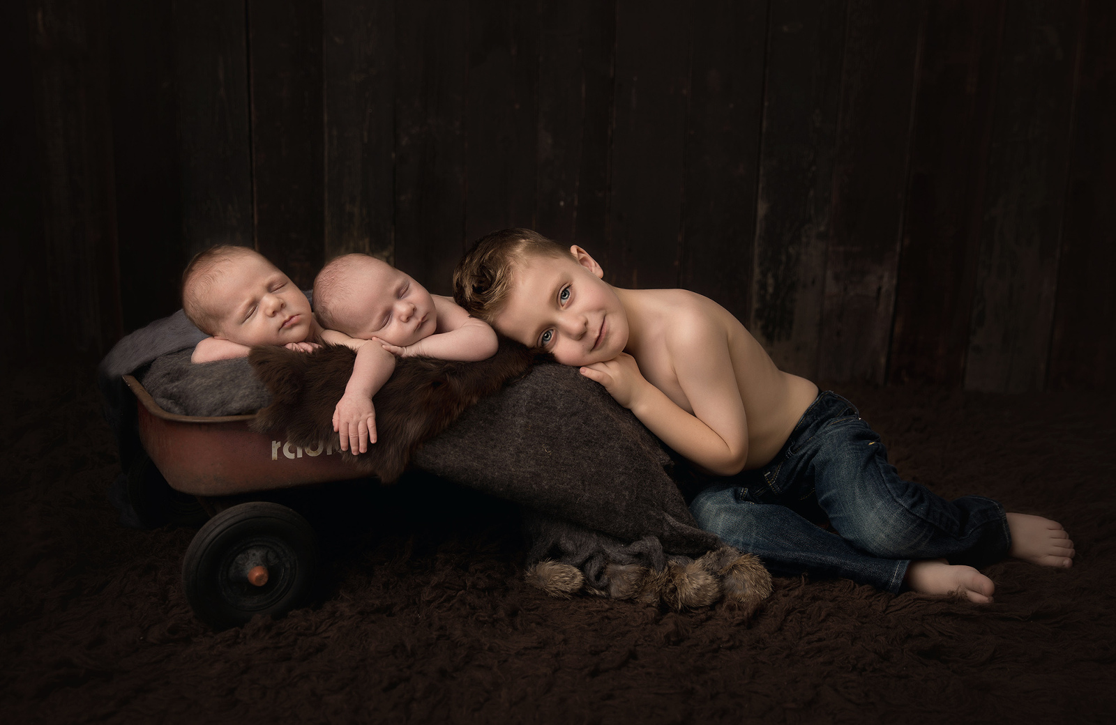 Studio newborn twins portrait session from the side with dark background of a proud older toddler brother lying down near his newborn twin siblings who are sleeping together on an antique wagon. Image by Helga Himer Photography, Sudbury, Ontario.