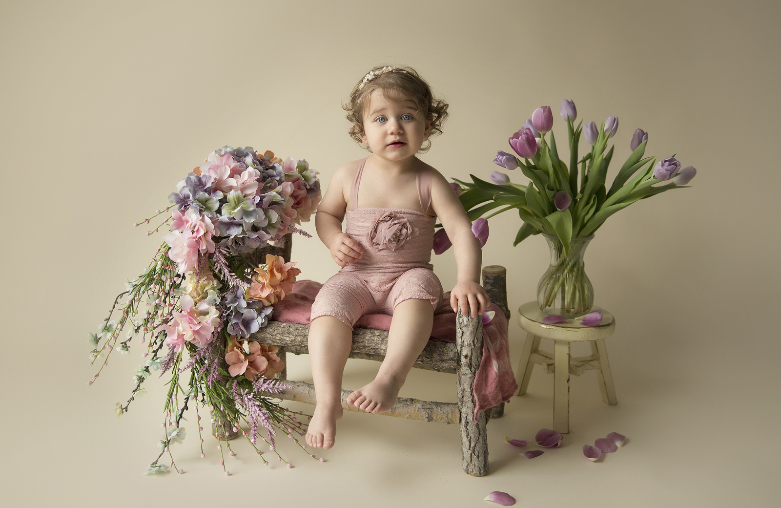 Studio child portrait photography session, a young blonde girl in pink jumper sitting on a custom-made wooden log bed, with flowers nearby, and a light background. Image by Helga Himer Photography, Sudbury, Ontario.